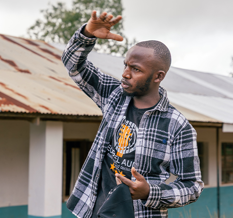 Eliud stands outside, making an expression with his hands as he speaks.