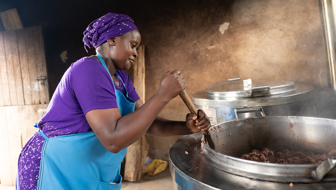 A woman in a a blue apron stirs a large pot over a stove.