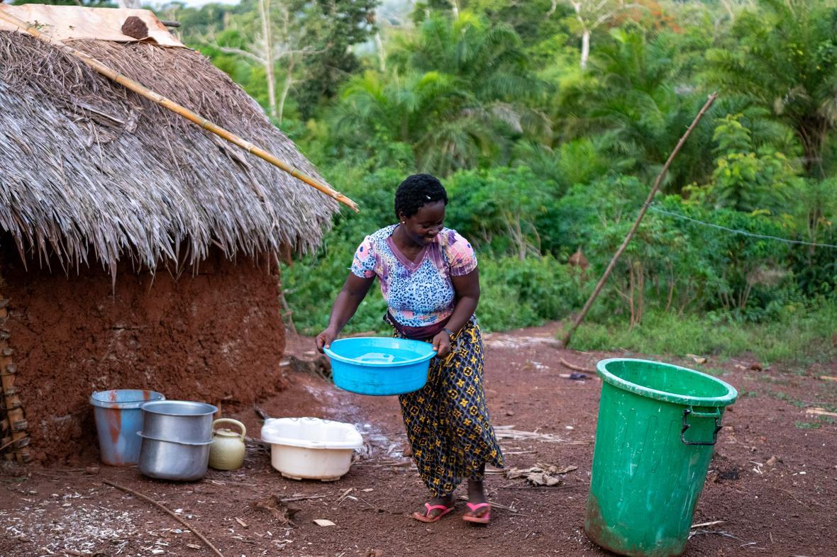 A woman carries a water bucket outside a home with lush rainforest in the background.