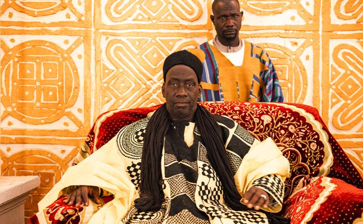 A king sits in his throne. Ornate patterns adorn his clothes, the red throne, and the wall behind.