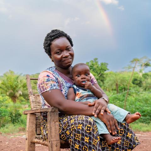A woman holds her child in her lap in a wooden chair. Green rainforest and a rainbow are in the background.