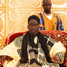 A king sits in his throne. Ornate patterns adorn his clothes, the red throne, and the wall behind.
